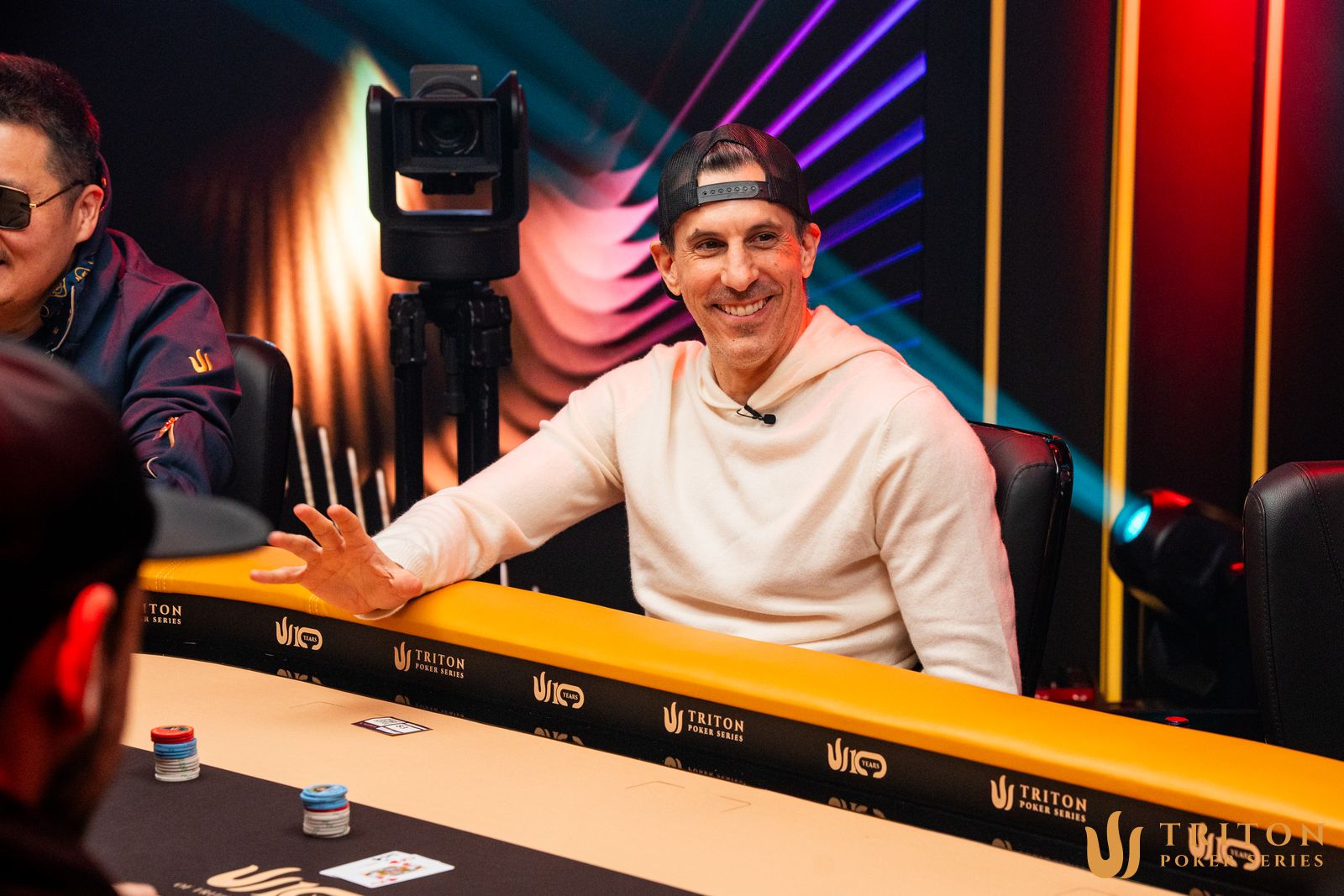 Zdjęcie stołu pokerowego podczas turnieju Triton Invitational z widocznymi żetonami i kartami.